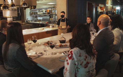 Group of people sitting around the Chef's Table with a server presenting a dish while wearing face mask