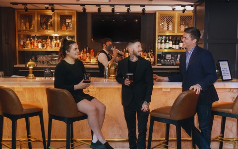 Three people talking at the bar with drinks in hand while bartender shakes cocktail