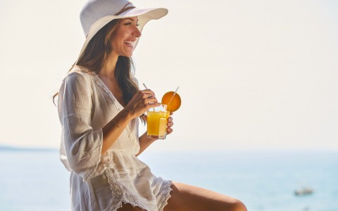 woman wearing a white dress and hat smiling while she is holding a glass of tropical drink