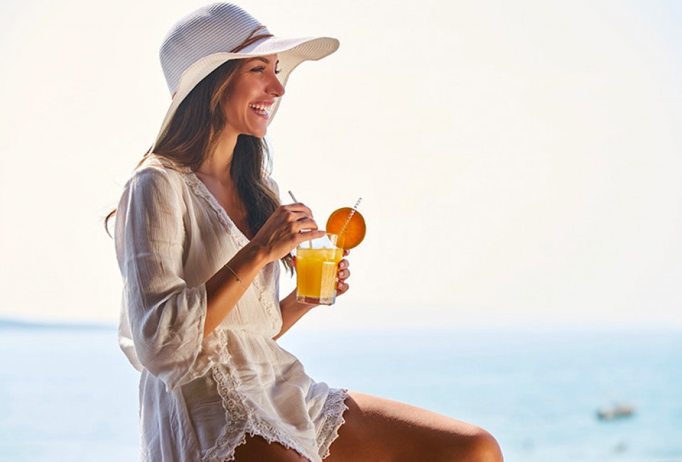 woman wearing a white dress and hat smiling while she is holding a glass of tropical drink