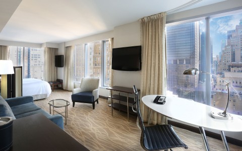 One bed room with large white desk and 3 very large windows looking over the city 