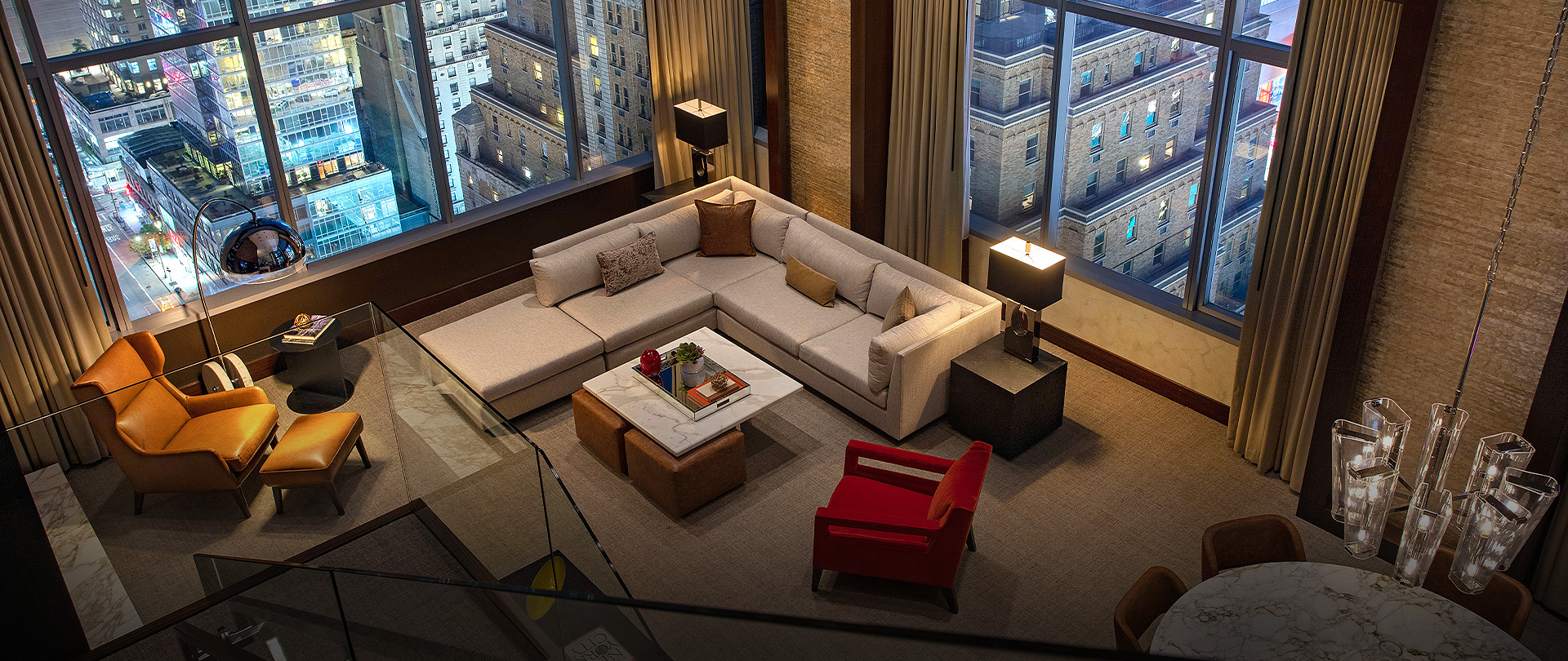 A light gray, beige l-shaped couch with brown pillows. floor to ceiling windows, and red accents in the suite