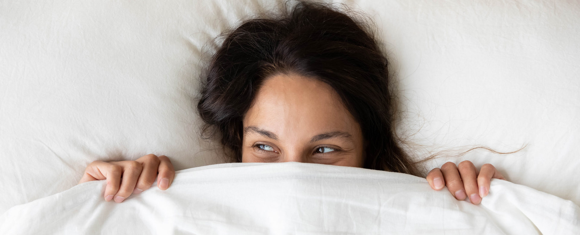 woman laying in bed holding white bedsheets just below her eyes