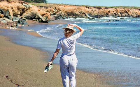 Back view of a woman holding her hat and walking down the beach