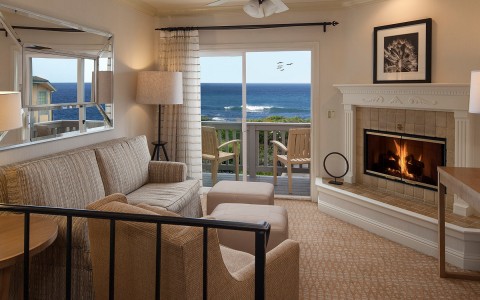 a living room with couches near a fireplace looking out a sliding glass door to a balcony with 2 chairs on it with a view of the ocean
