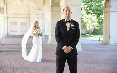 groom standing in black suit with bride walking up behind him in long white gown 
