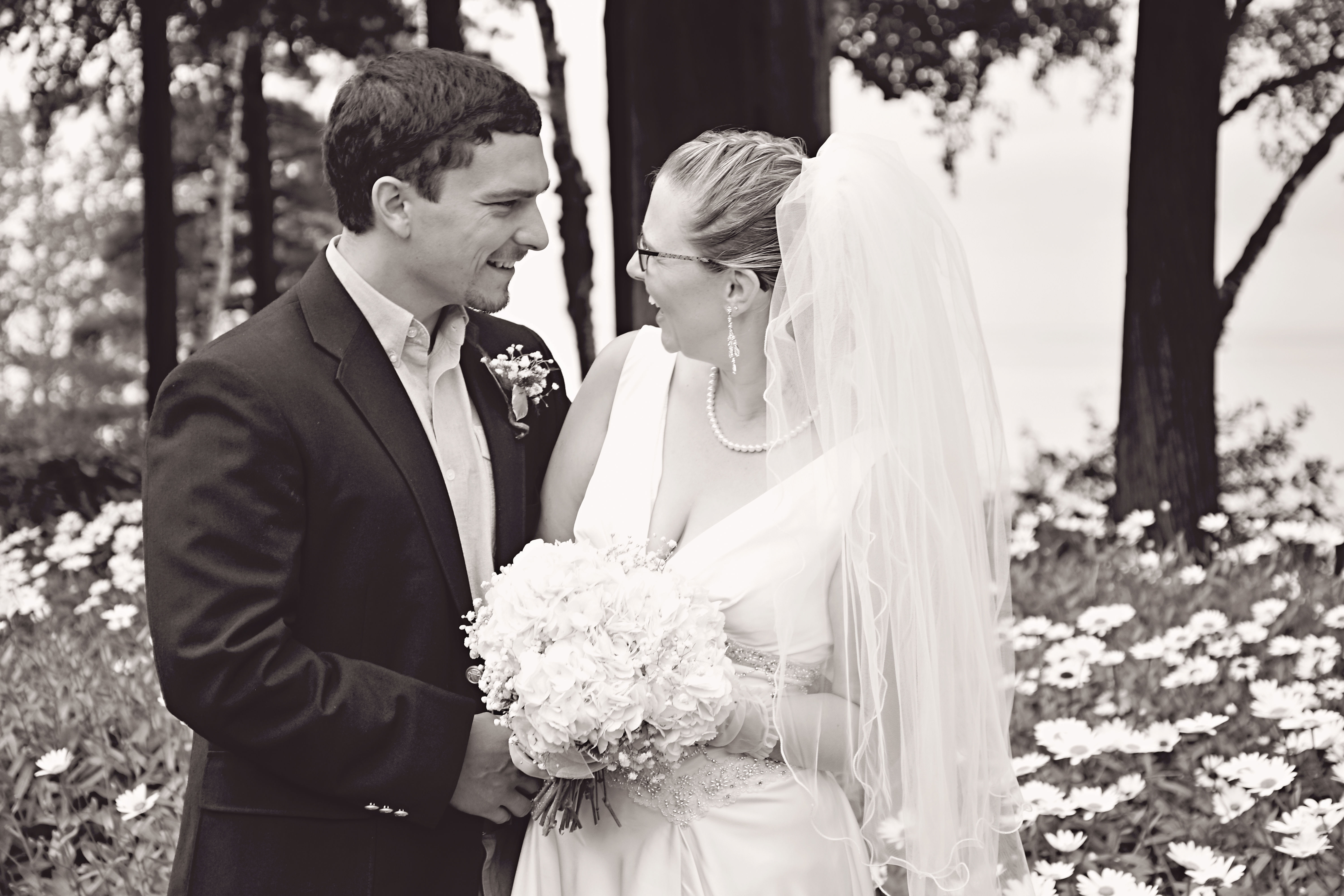 Black and white photo of groom and bride smiling at each other with garden and flowers in the background