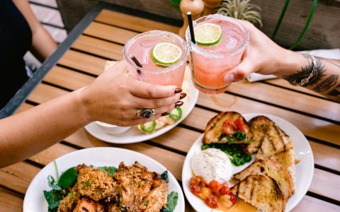 male and female hands making a toast with a watermelon margarita on top of their meals served on the table