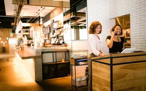 two restaurant hostess standing up smiling while one of them is taking a phone call