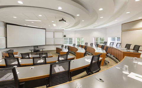Arced desks with several seats tied down towards a desk with a large projector behind it