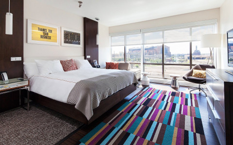 A bed is near a long window with a view of the city and a long artistic rug of various cool tones sits at the foot of the bed