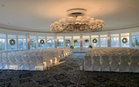 A large ballroom decorated for a wedding ceremony with large windows lining the walls overlooking a garden