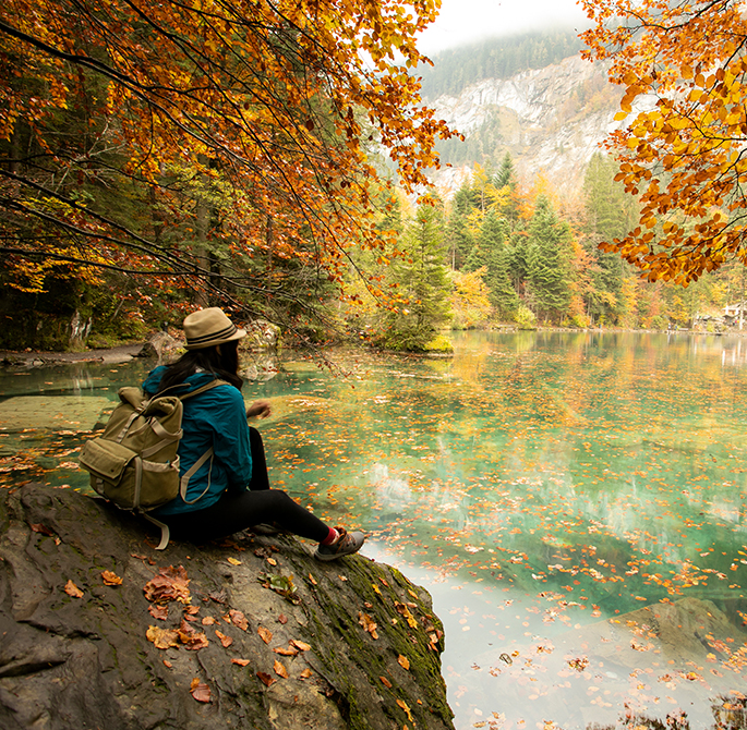 a woman in fall clothing sitting on a large boulder overlooking a lake with fall foliage overhead