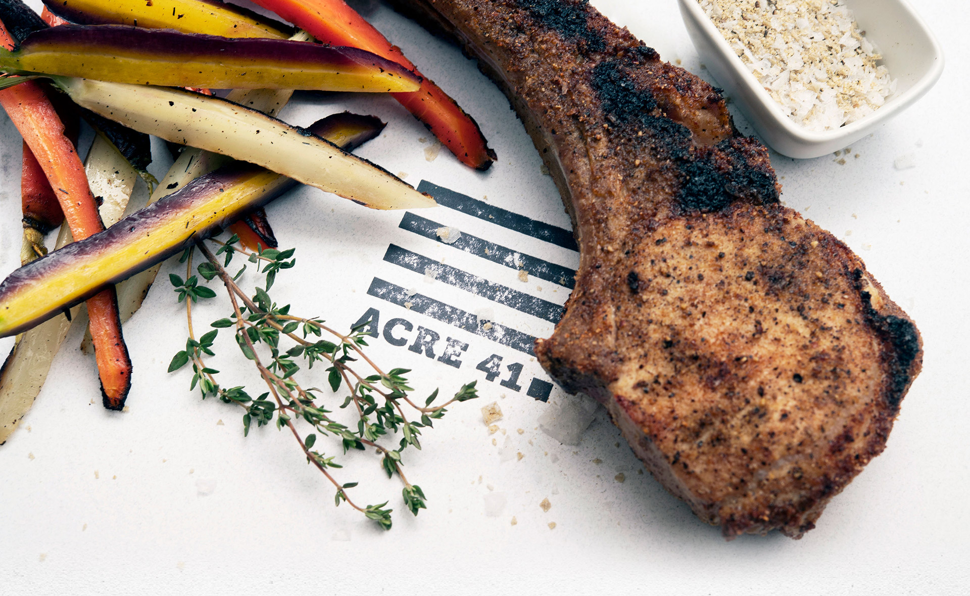 a pork chop with colorful sticks of potatoes and veggues on a white table with the acre 41 stamp on it