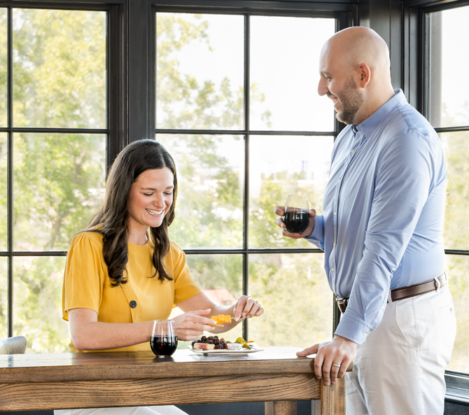woman grabbing a piece of cheese while a man is standing by her side with a glass of wine