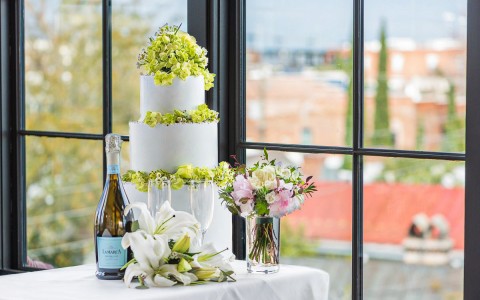 view of a wedding cake with flowers and a bottle of champagne with a window behing