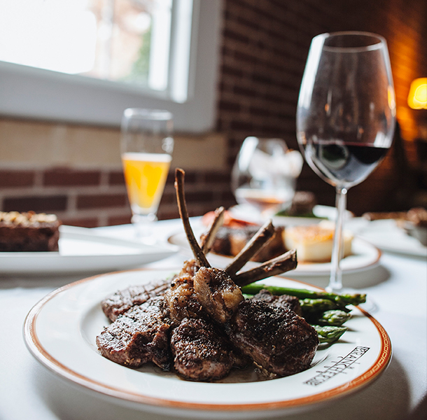 close up of lamb chops with asparagus with a glass of red wine and other dishes in the background