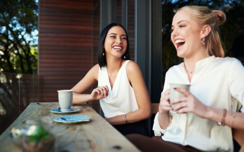 two women sitting and drinking coffee while laughing
