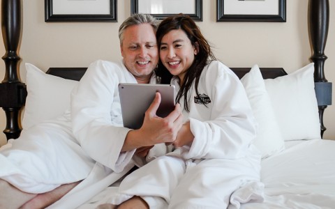 a couple sitting in bed with their robes on while holding a tablet 