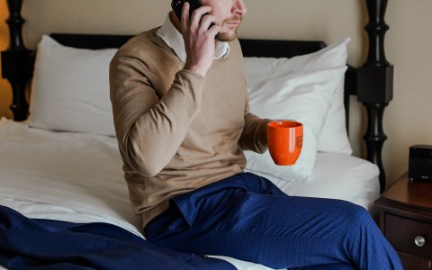 man sitting on a bed while holding an orange mug and holding a phone to his ear 