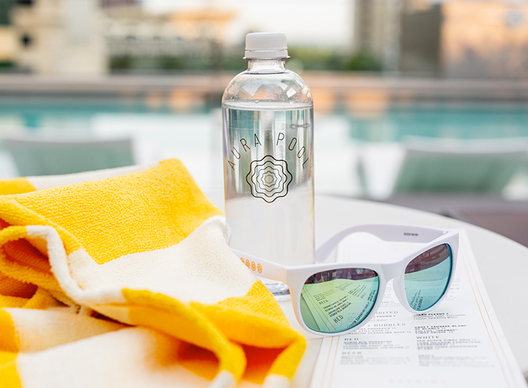 aura water bottle and sunglasses with yellow and white towel with the pool in the background
