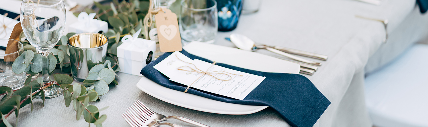 close up shot of white plate with blue napkin and white menu tied together with string and green plants in the middle of the table