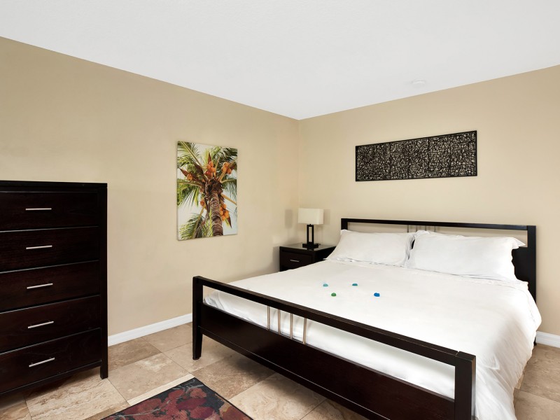 white linen bed, long black and white picture, nightstand to the left, dresser in left corner and palm tree picture on wall
