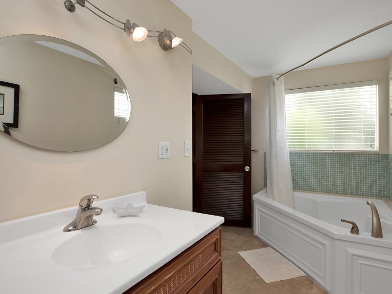 white tiled bathtub/shower, white counter sink w circular mirror above it and wood door leading outward