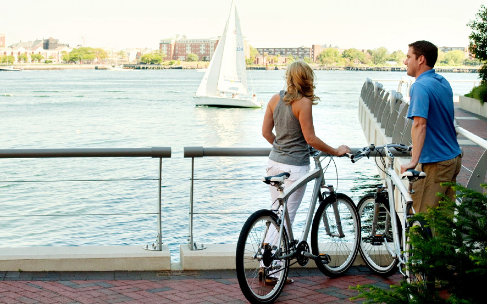 man and woman with bikes looking out to the wharf and a sailboat