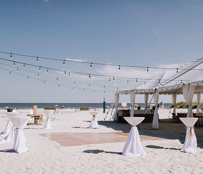 many high top tables on the sand with strings of lights hanging over them