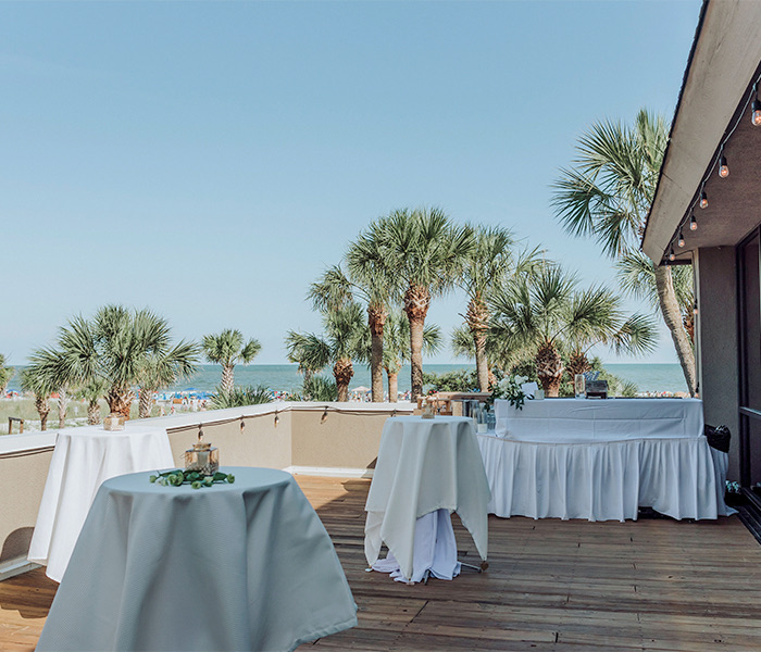 a balcony with high top tables and a bar overlooking the ocean and palm trees