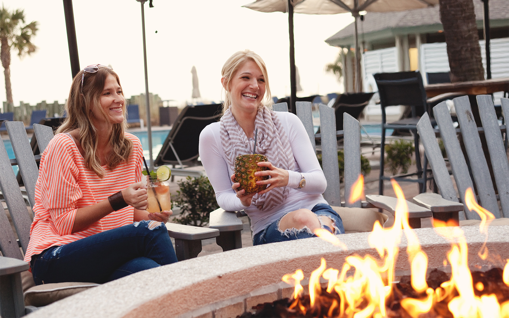 two women smiling and holding drinks while sitting around a lit fire