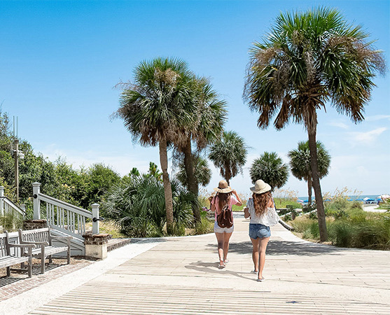 two girls walking on a path with benches to the left and the beach to the right