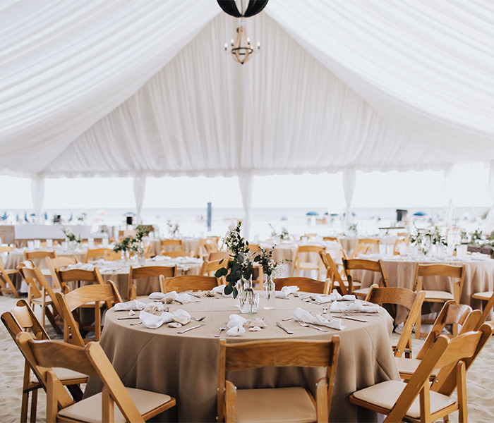 many tables with light brown linen and light wood chairs under a white tent with a chandelier hanging from the middle