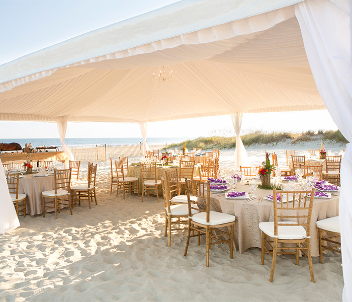 outdoor tables and chairs under a large white tent on the beach with the ocean in the distance