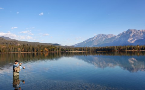 Man Fishing in Lake with Trees and Mountains in Background