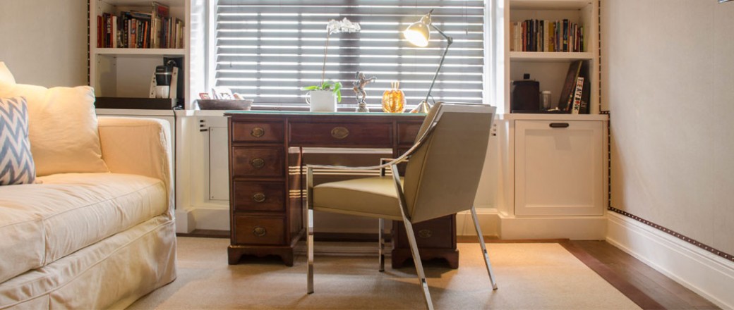 Beige sofa next to wooden desk with chair & book shelf in the back