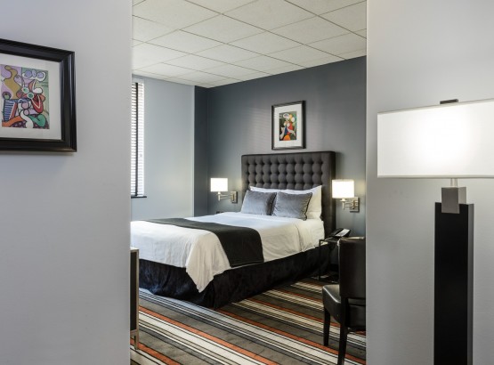 Entryway into room with art hanging on left wall, standing lamp on right side, queen bed directly in front with black headboard, black throw at the foot, white comforter and sheets, grey decorative pillows