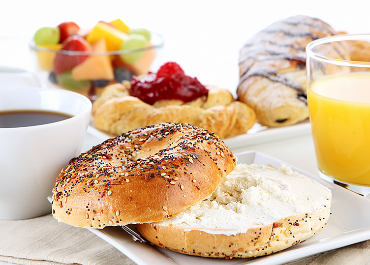 An everything bagel, a cup of coffee, a cup of orange juice, a croissant and a bowl of fruit on a table