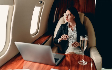 A woman sitting in an airplane wearing a black blazer and looking out the window with a laptop and glass of champagne in the front of her on a table.