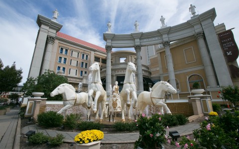 A large white statue of horses and tall columns behind with lots of plants to the right.