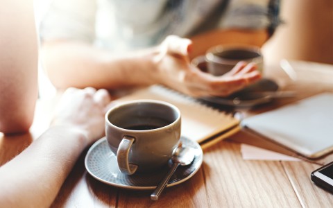 Two people enjoying a cup of coffee with notebooks and the sun shining in from the background.