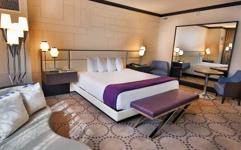 A bedroom with a large bed and lots of low lit lamps as well as a large floor mirror.