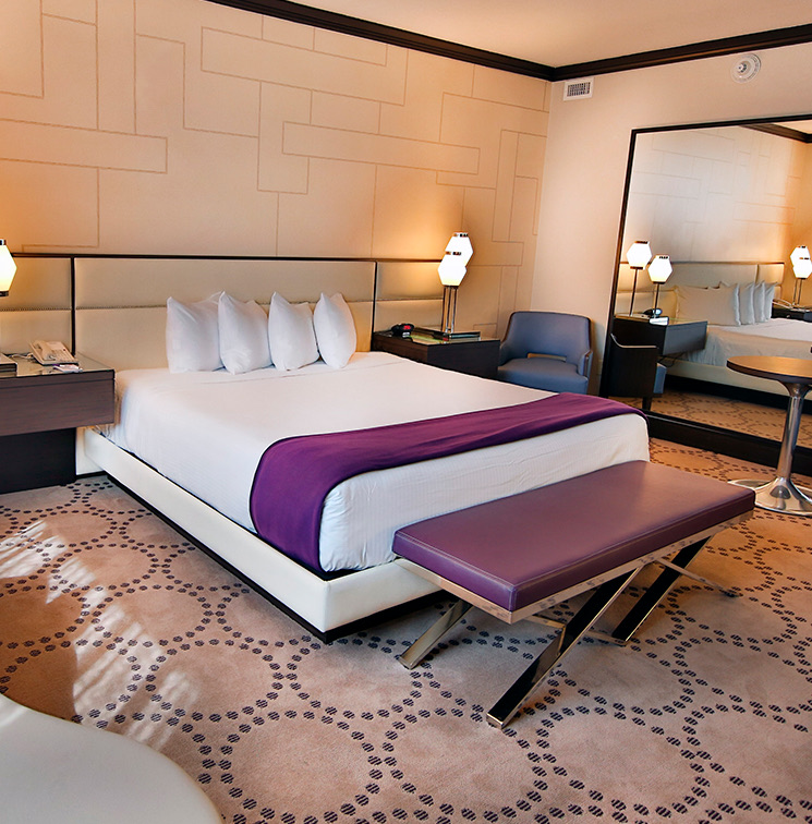 A large bed with purple sheets and bench in front and low lit lamps to each side.
