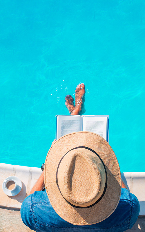 A person wearing a fedora hat reading a book with their feet in the pool.