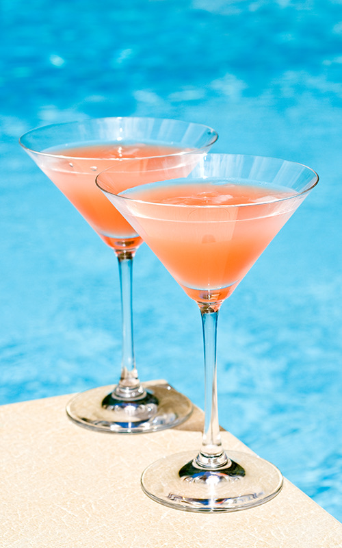 Two pink drinks served in a martini glass on the deck next to the pool.