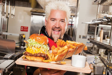 Guy fieri holding a wooden platter of fries and a cheeseburger.