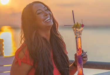 A smiling woman holding a tall, skinny cocktail with the sun setting in the distance.