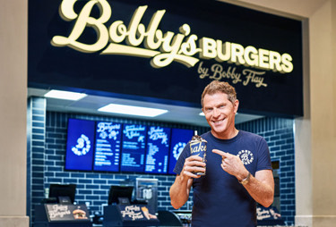 Bobby flay smiling and pointing to a milkshake in his hand while standing in front of Bobby's burgers.