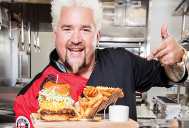 Smiling guy fieri pointing to a cheeseburger and fries.